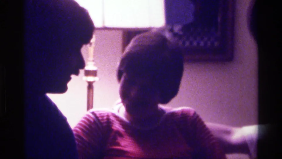 A man in profile faces a seated woman in a softly lit 1976 Kentucky home interior with a lamp and wall picture behind them.