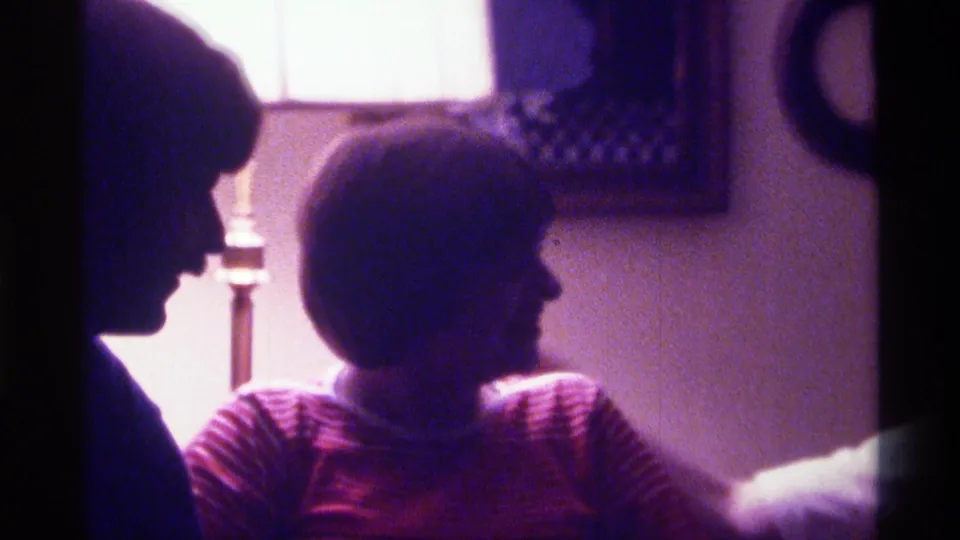 The man and seated woman appear turned toward one another in a grainy 1976 Kentucky home-movie interior.
