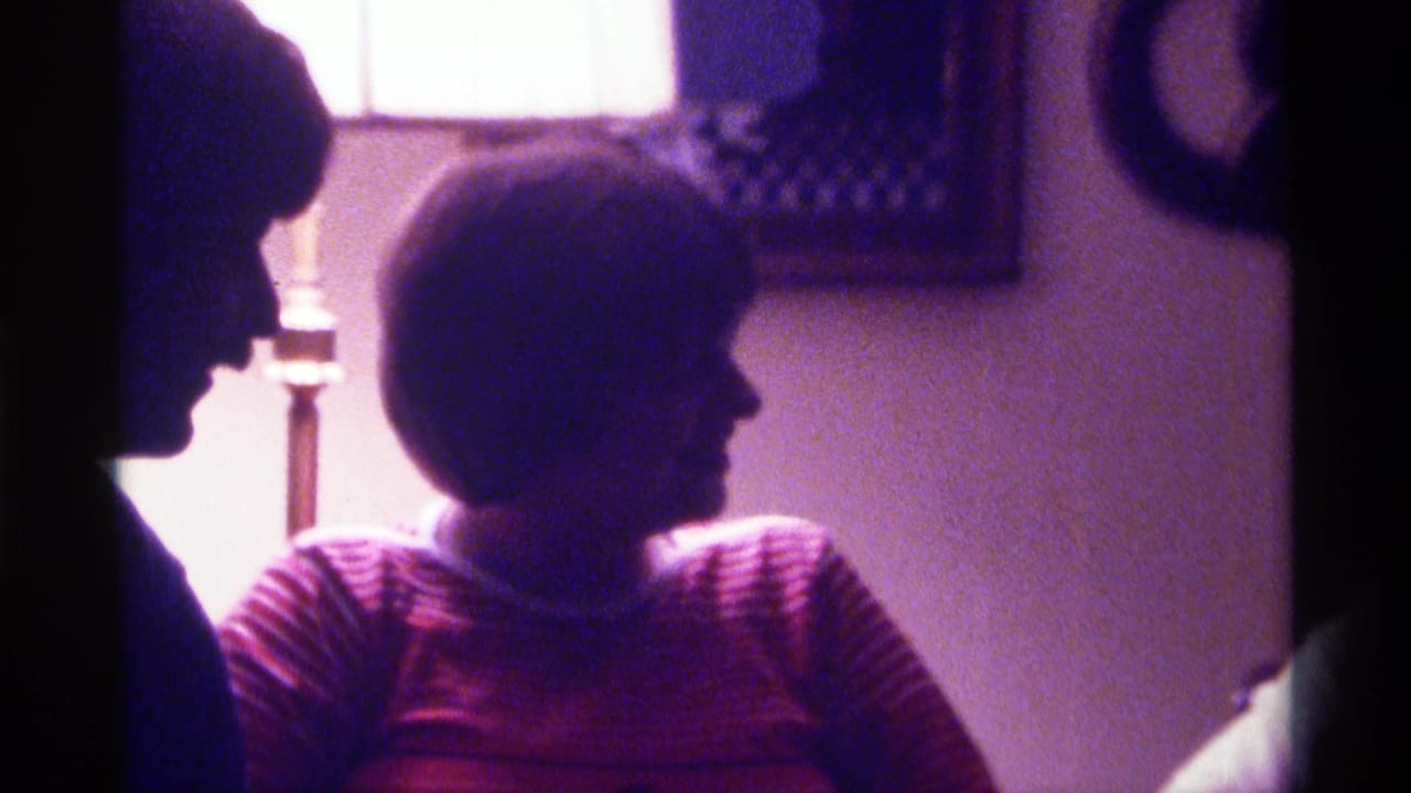 1976 Kentucky home-movie frame showing adults in conversation indoors, including a smiling woman in a striped sweater near a lit table lamp.