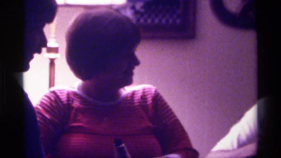 1976 Kentucky home-movie frame with a woman in a striped sweater seated in profile while another adult silhouette faces her indoors.