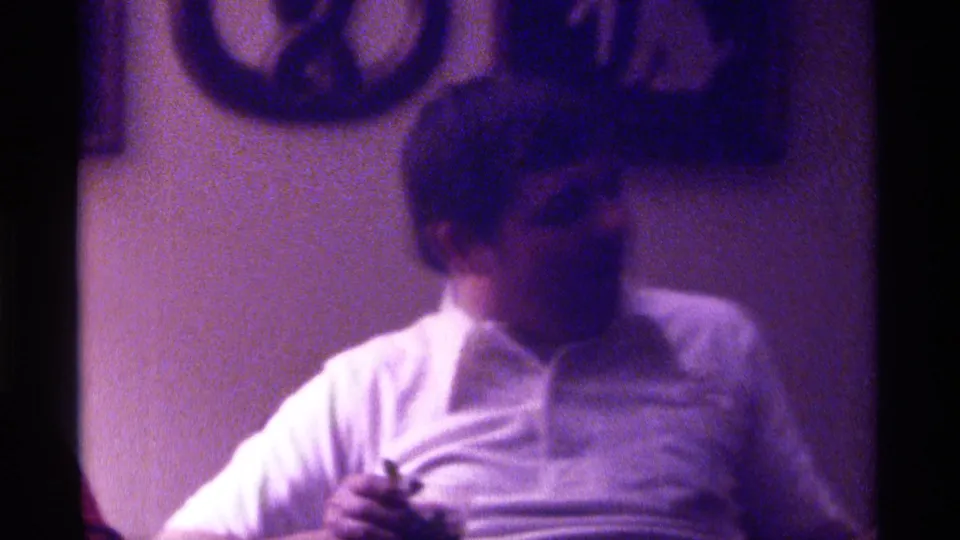 1976 Kentucky home-movie frame showing a seated man in a white shirt in a dim living room, with soft focus and wall decor behind him.