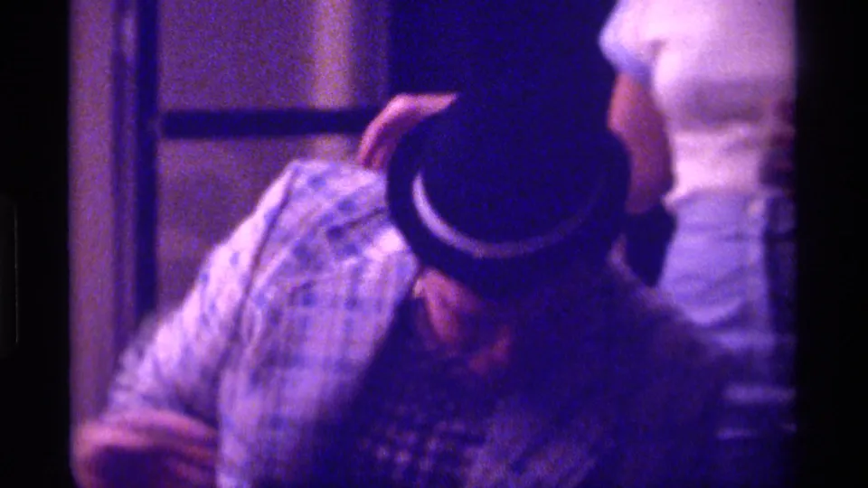 1976 Kentucky home-movie frame showing a seated man in a patterned shirt and brimmed hat within a softly lit private living-room scene.