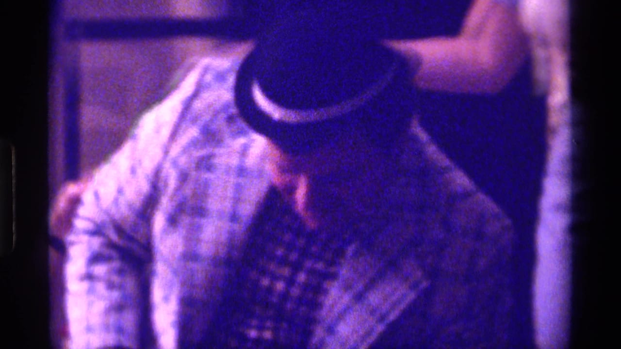 1976 Kentucky 8mm frame of a seated man in a plaid jacket and small dark hat, leaning forward in a tan upholstered chair.