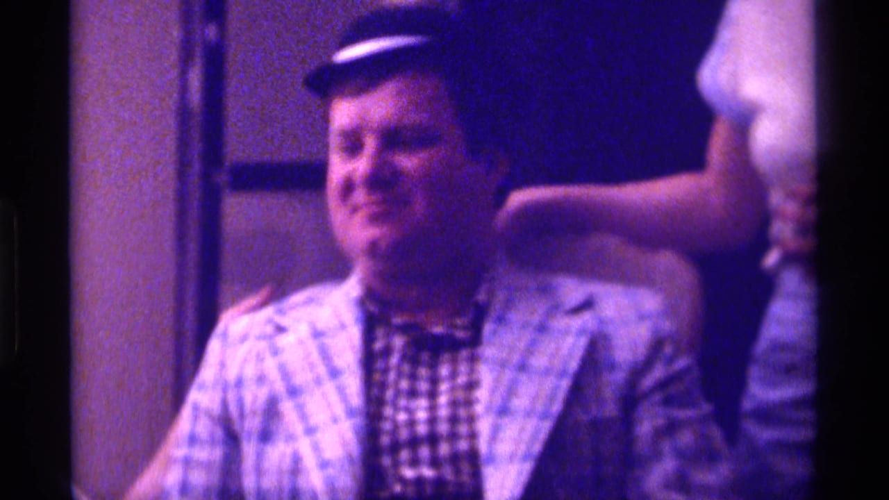 Man in a small dark hat sits in a tan chair with a hand resting on his shoulder.