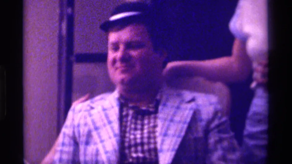 1976 Kentucky home-movie frame showing a man in a plaid jacket seated in a tan chair as a nearby hand rests on his shoulder.