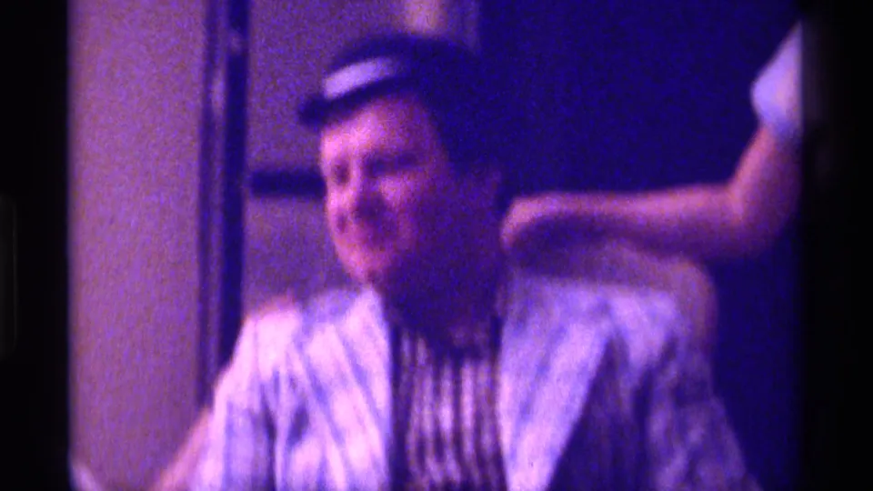 Blurred 1976 Kentucky 8mm frame of the man seated indoors in a plaid jacket and small dark hat, with the chair and wall behind him.