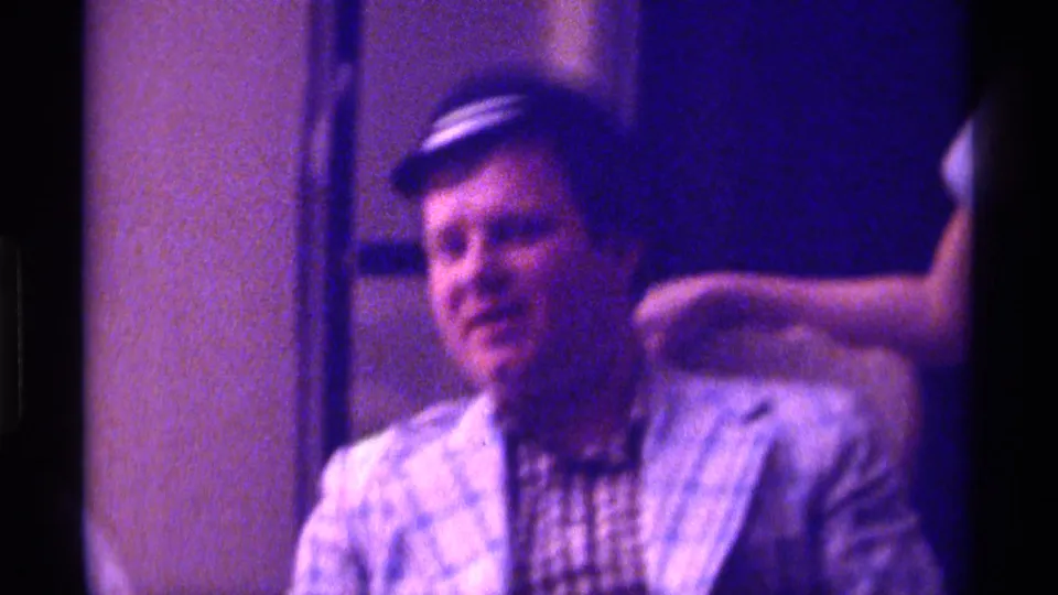 1976 Kentucky 8mm frame of the seated man in a plaid jacket and small dark hat, with another person's arm and hand near his shoulder.