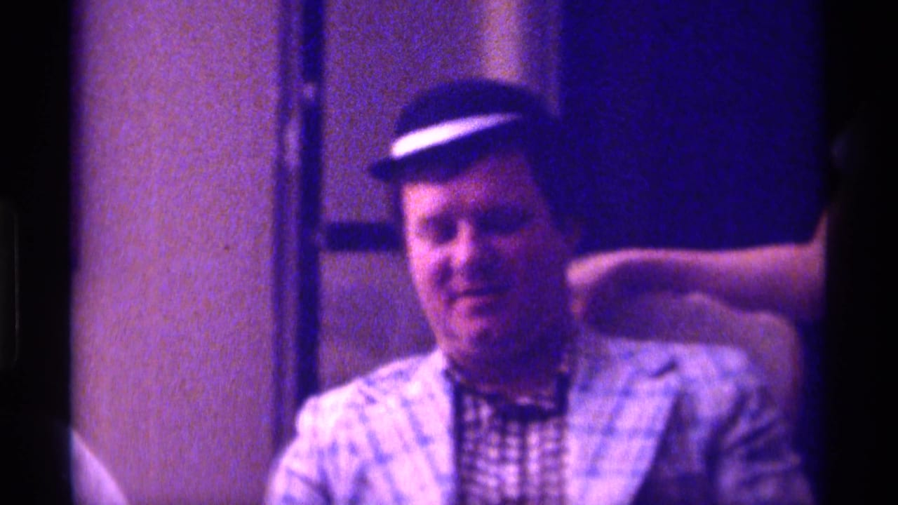 Seated man in small black hat and plaid jacket indoors under purple-tinted light.