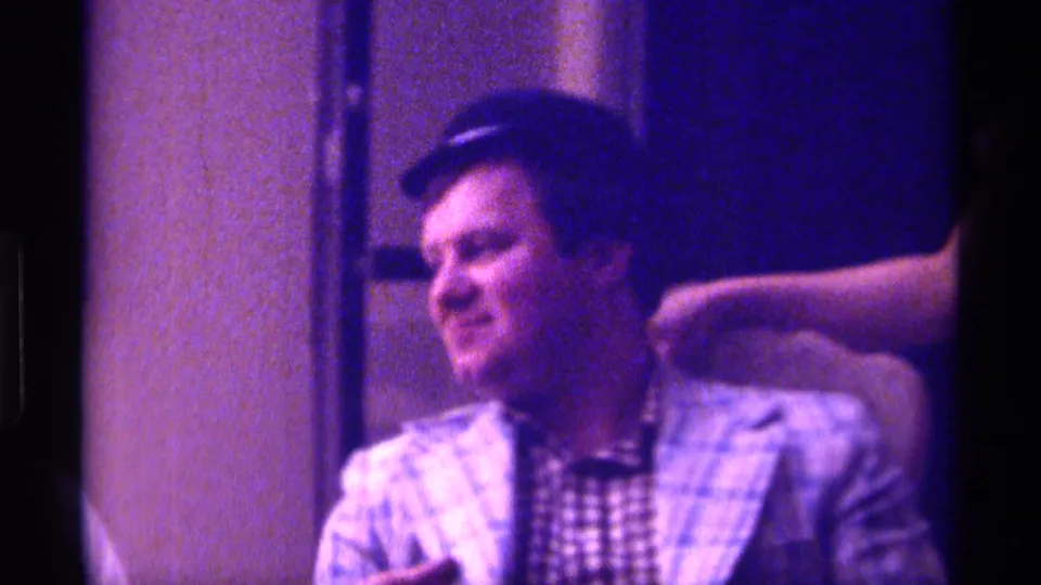 The adult man in the small dark hat turns to the side in the dim 1976 Kentucky home-movie interior, suggesting a candid exchange off camera.