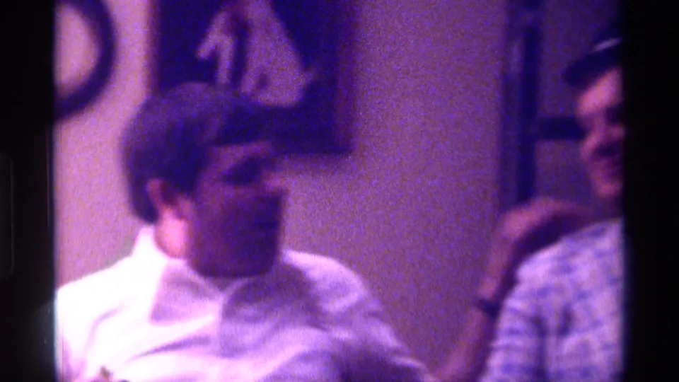 A second adult man in a white shirt appears in profile inside the 1976 Kentucky home-movie scene, with the plaid-jacketed man partly visible nearby.