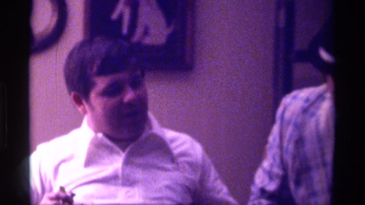 Adult man in a white collared shirt sits or leans indoors beside another person.