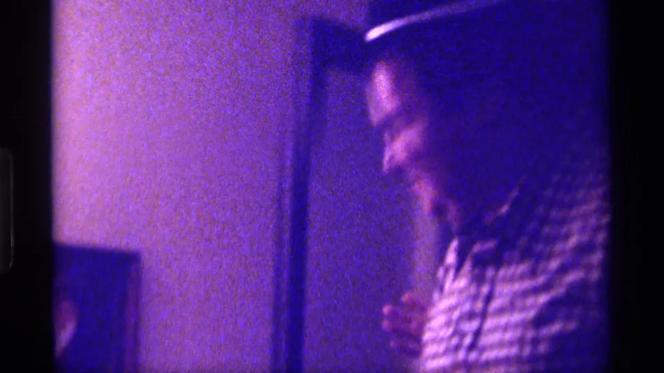 The adult man turns in profile while wearing a dark hat and checkered shirt in the grainy 1976 Kentucky interior footage.