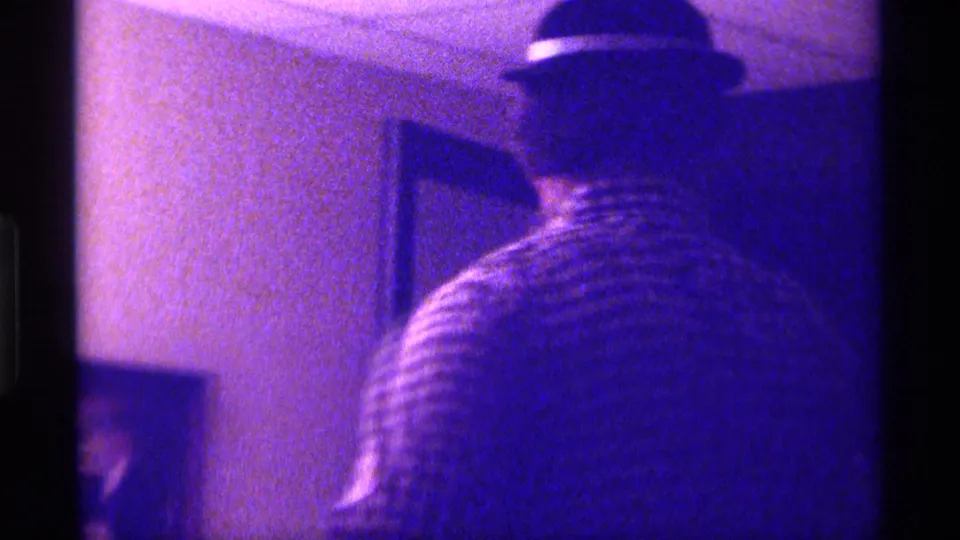 The man is seen from behind in a dark hat and checkered shirt as he faces a wall or doorway inside the private Kentucky interior.
