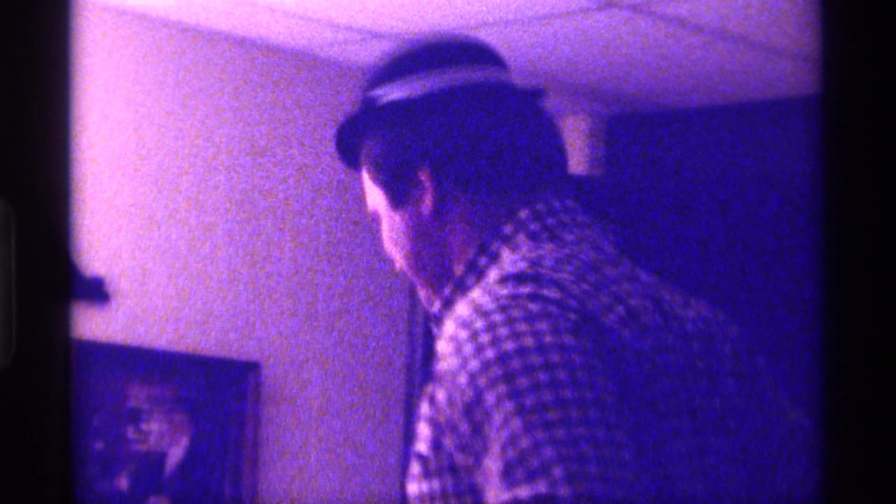 Man in a checkered shirt and brimmed hat turns inside a dim room.