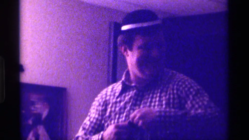 A man in a checkered shirt and brimmed hat faces the camera area while holding or arranging clothing in a Kentucky home interior.