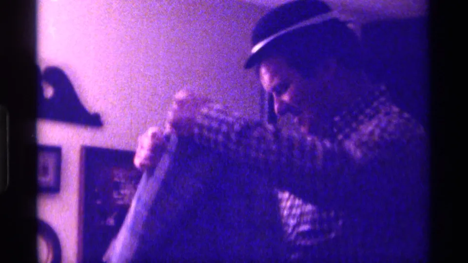 The man leans in to help a child with a jacket or garment inside the dim 1976 Kentucky home-movie scene.