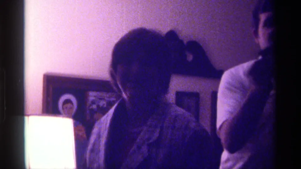 A woman in the winter coat appears to smile toward the camera as a man stands beside her in the softly lit 1976 Kentucky home movie.