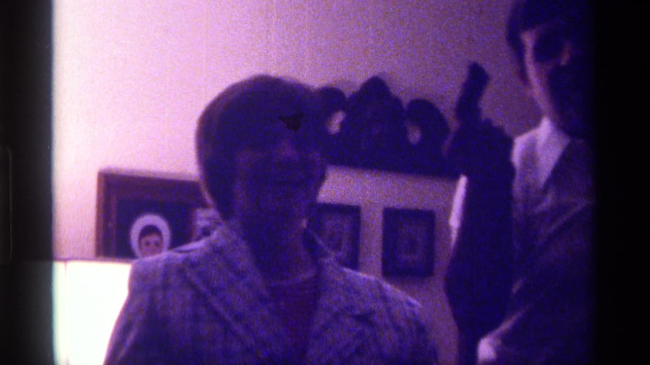Woman seated indoors with a young man nearby, framed photos and a shelf behind them.
