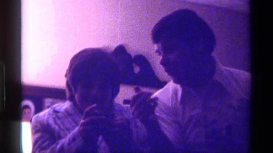 The woman and young man sit close in the living space, likely looking at a photograph or small item, with framed pictures behind them.