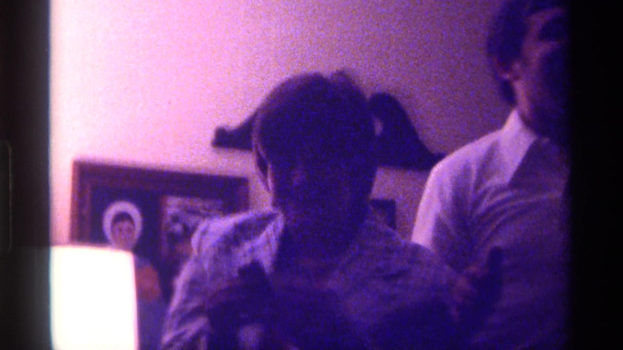 Two people in a dim living room near framed wall photographs and a bright lamp.