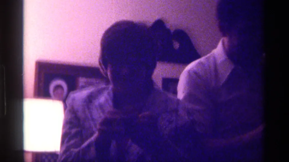 1976 Kentucky home-movie frame with a seated figure, another person nearby, framed wall decor, and a bright lamp in purple-tinted light.