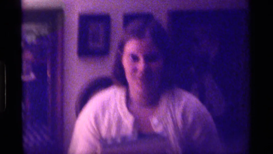 1976 Kentucky 8mm frame showing a person seated indoors before framed pictures, with soft focus, grain, and purple color shift.
