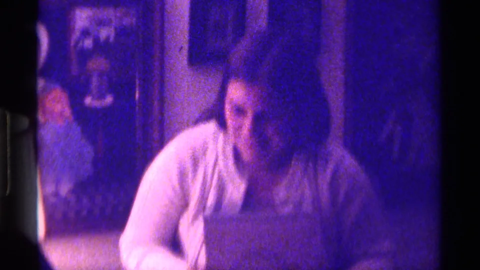 1976 Kentucky home-movie frame with a seated person looking down at a small object as another figure stands nearby in a private living room.