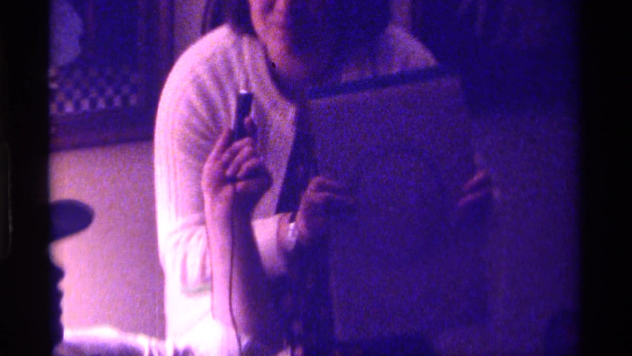 Woman in a white sweater holds a notebook and small pen-like object in a dim room.