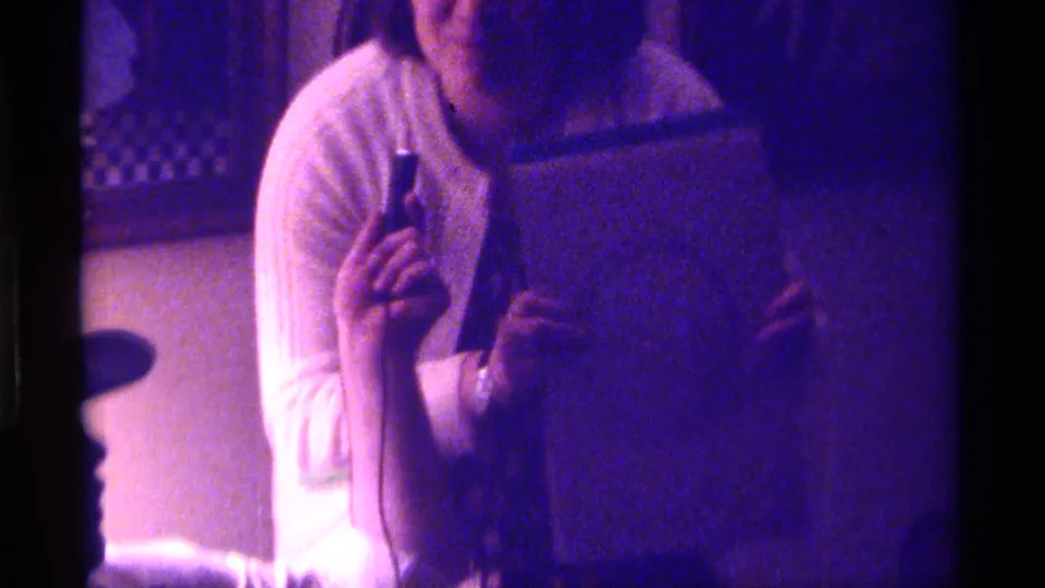 In 1976 Kentucky home-movie footage, a woman in a white sweater holds a notebook and pen-like object while seated indoors.