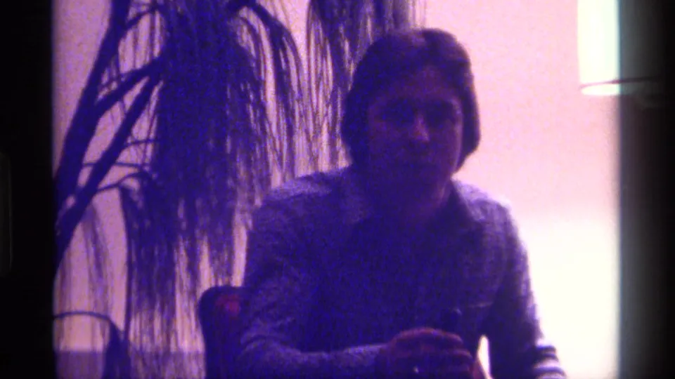 A seated man in a patterned shirt appears beside a tall houseplant in the purple-tinted 1976 Kentucky home movie.