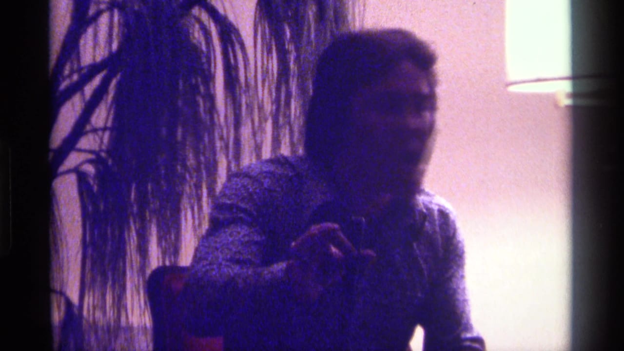 In Kentucky 1976 home-movie footage, a seated adult man holds a microphone near a plant and bright lamp in a dim interior.