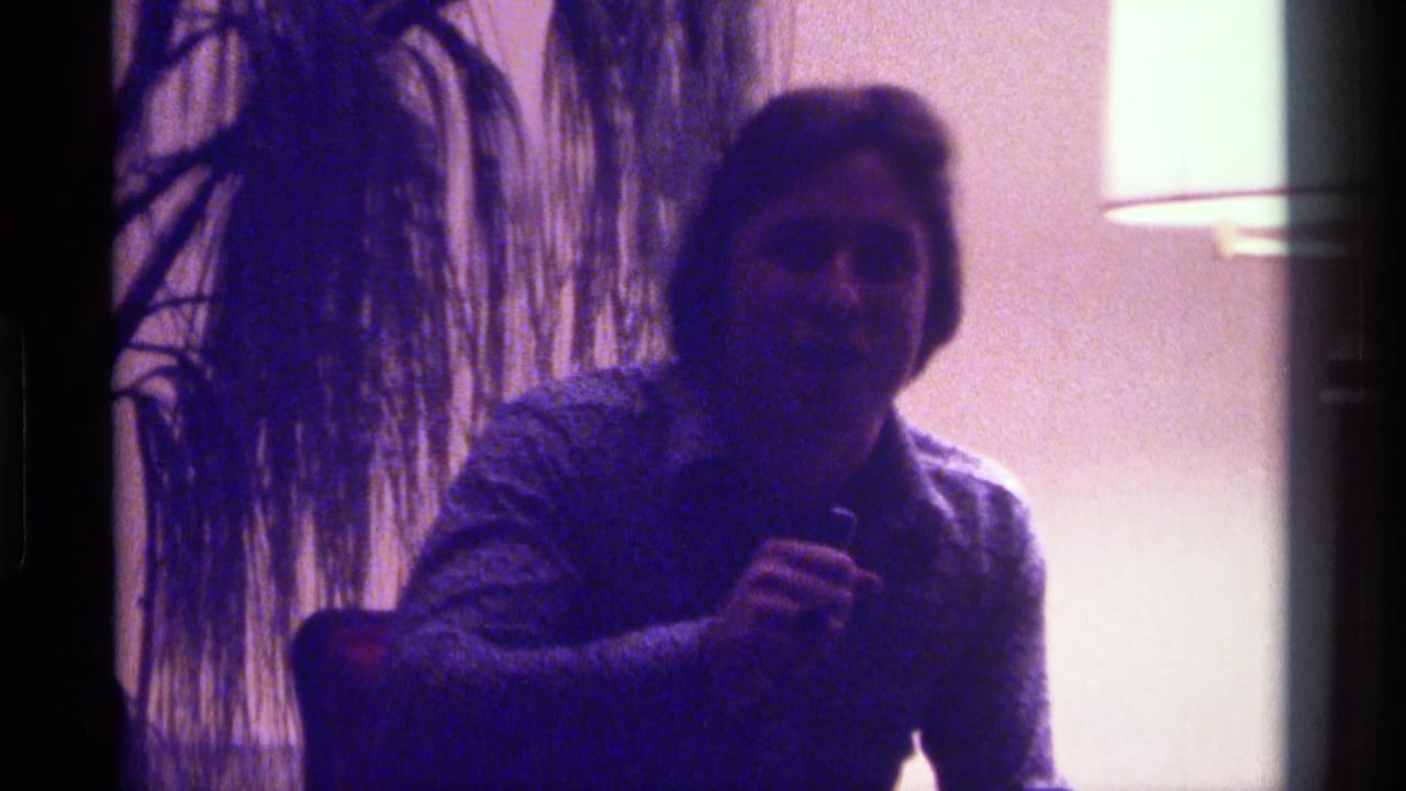 Man faces the camera while holding a microphone, with a plant and glowing lamp in the background.