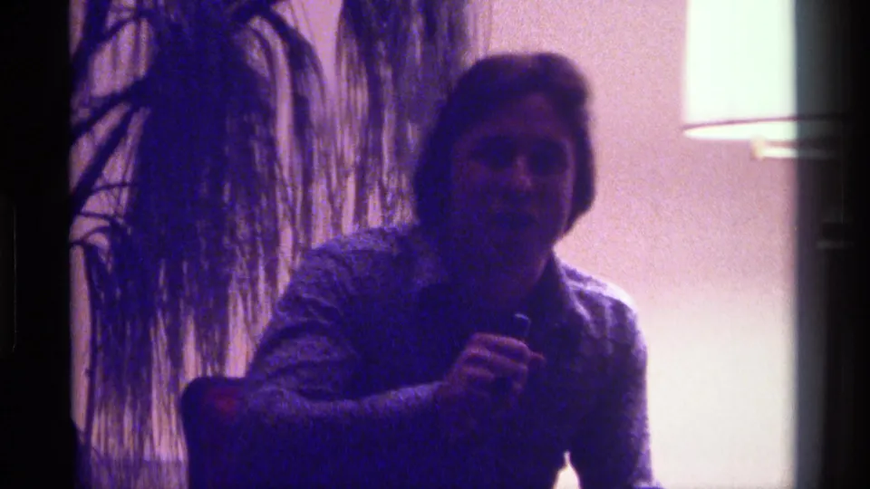 The seated man faces forward with a microphone during a Kentucky 1976 home gathering, framed by a plant and bright lamp.