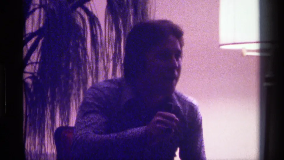 A seated adult man in Kentucky 1976 home-movie footage holds a microphone while turning slightly in a softly lit room.