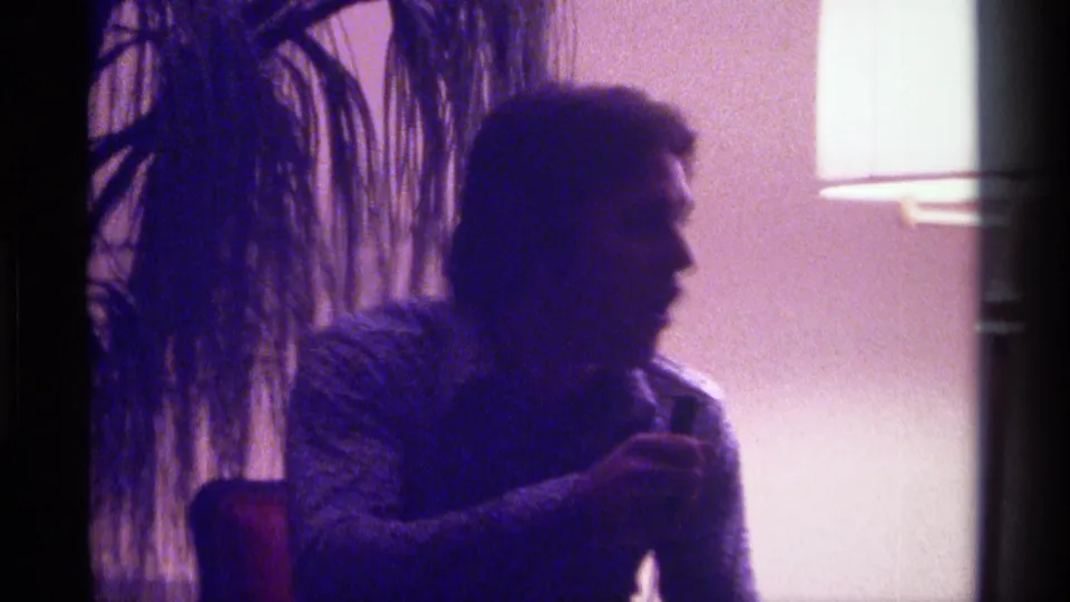 The performer turns into profile with the microphone in hand, still seated in the Kentucky 1976 home interior.