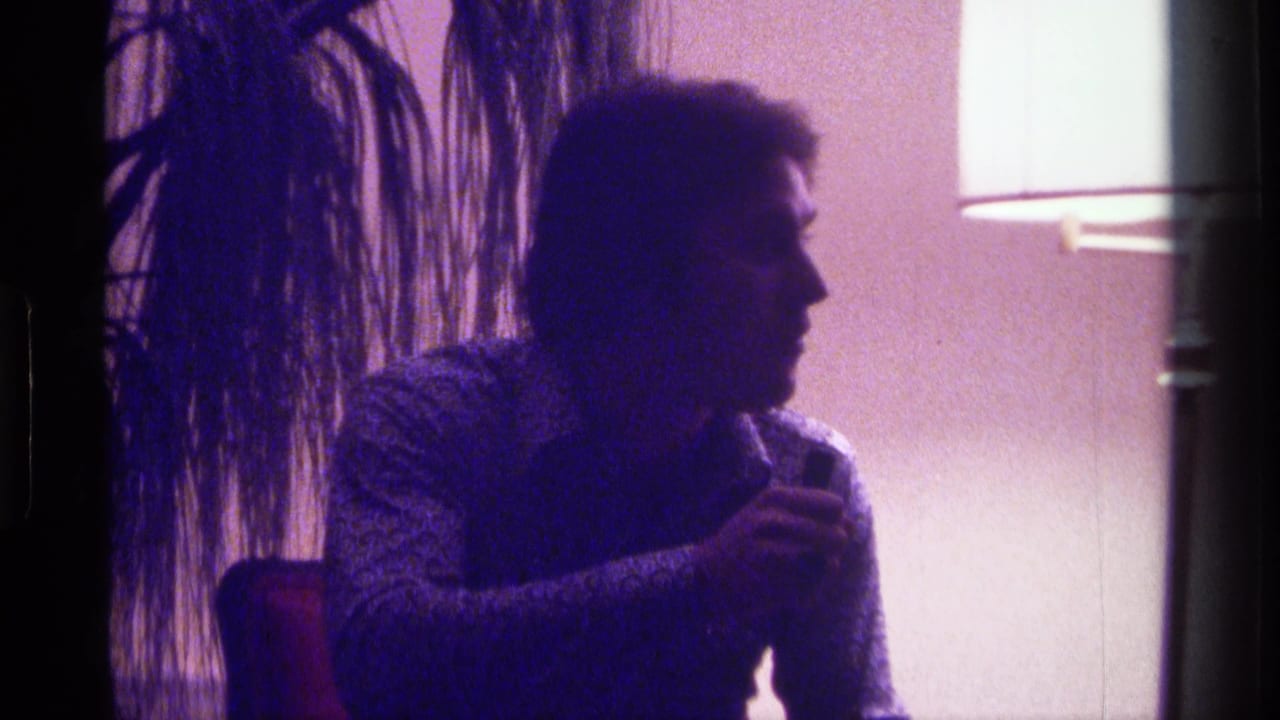 Man in profile sits indoors near a lamp and potted plant, holding a cigarette.
