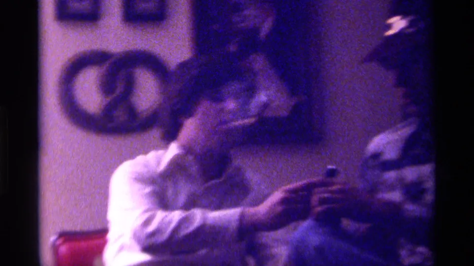 An adult seated in a Kentucky living room holds a cigarette near another person, with framed wall art visible behind them.