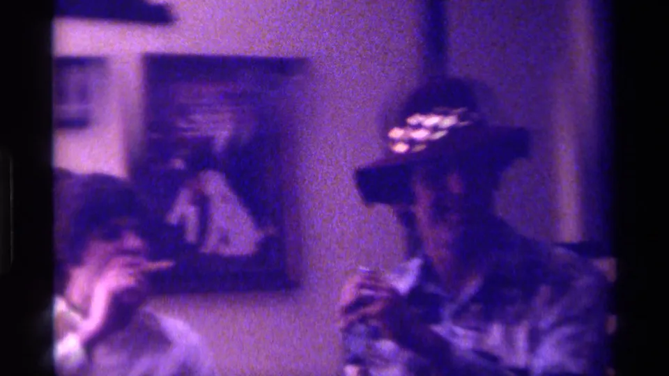 Two adults sit in a dim Kentucky home interior, one holding a cigarette, with framed pictures and wall decor in the background.