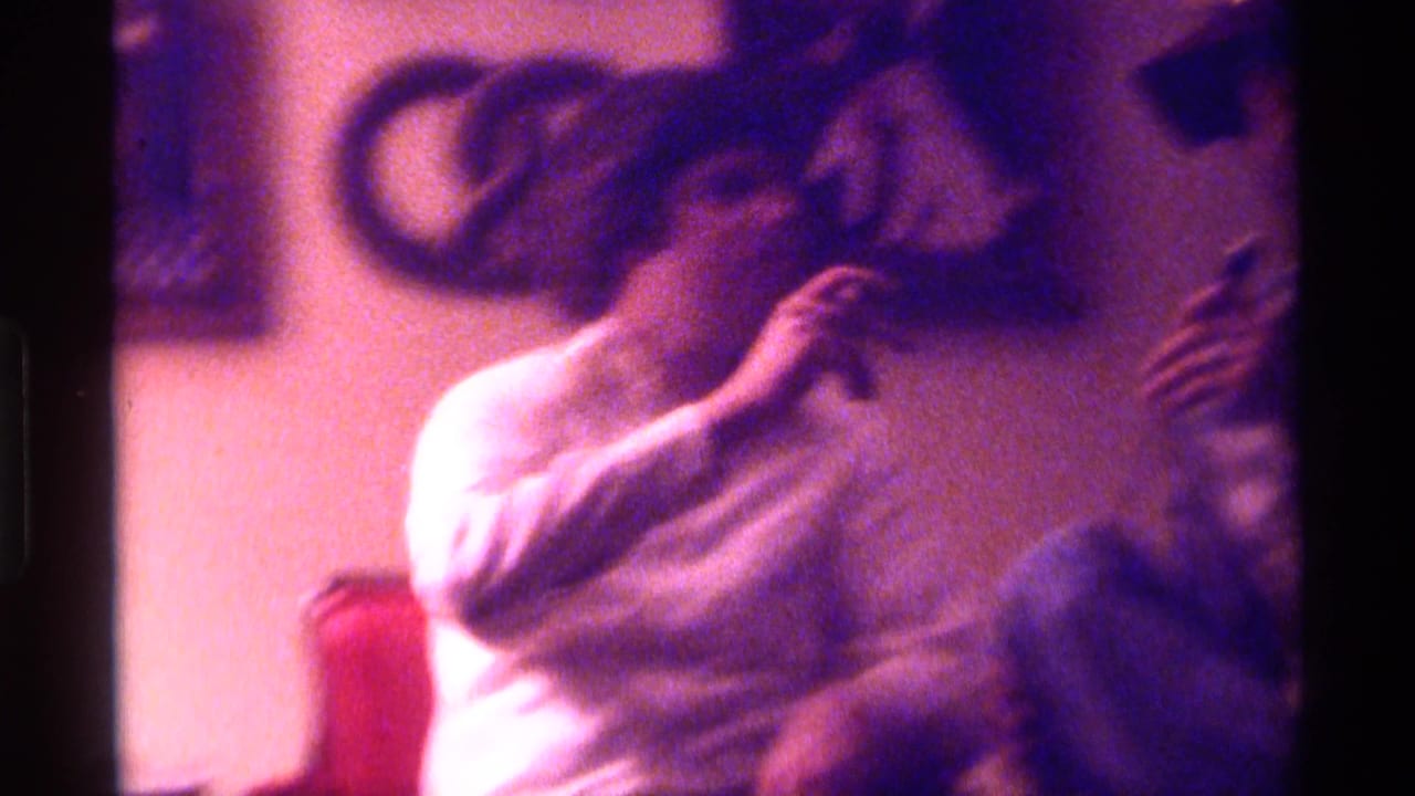 Two men seated indoors, one in a white shirt smoking while another figure is partly visible beside him.