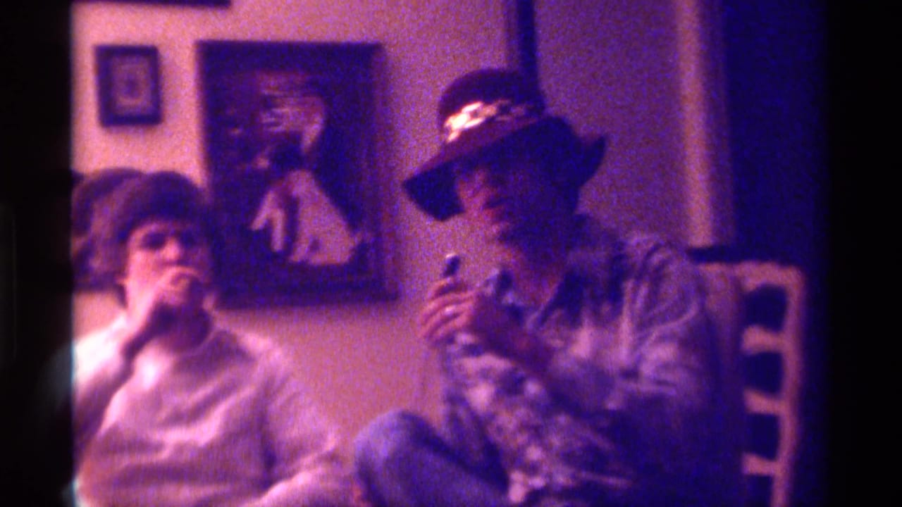 Two seated men smoke in a living room, with the man on the right wearing a brimmed hat.