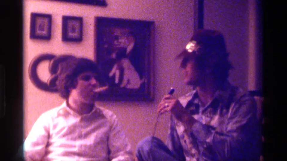 1976 Kentucky 8mm frame of two seated men in a living room, with cigars, smoke, a hat, and framed artwork behind them.