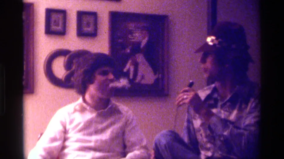 1976 Kentucky home-movie frame of two adult men smoking cigars in a private living room with framed art and circular wall decor.
