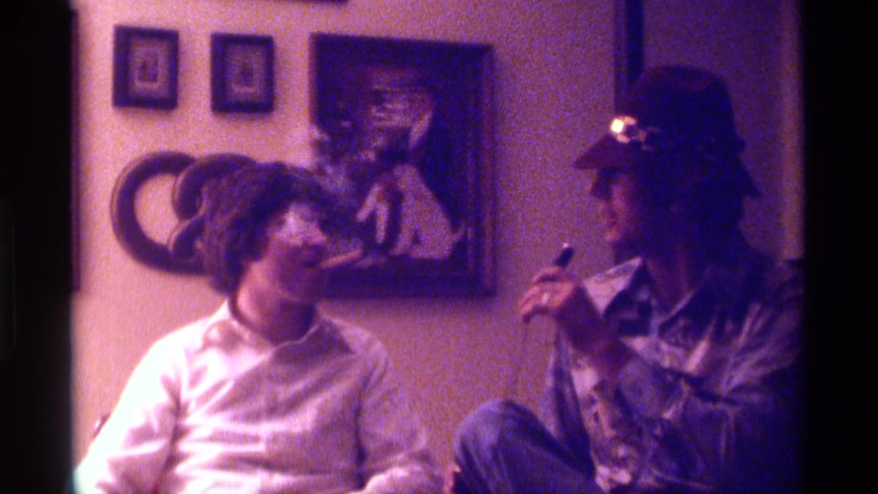 Two young men sit indoors, one holding a microphone toward the other in front of framed wall art.