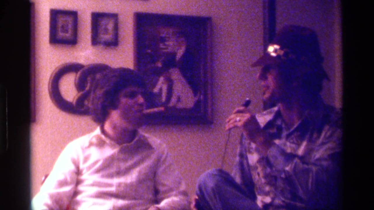 A young man in a hat holds a microphone while facing another seated young man indoors.
