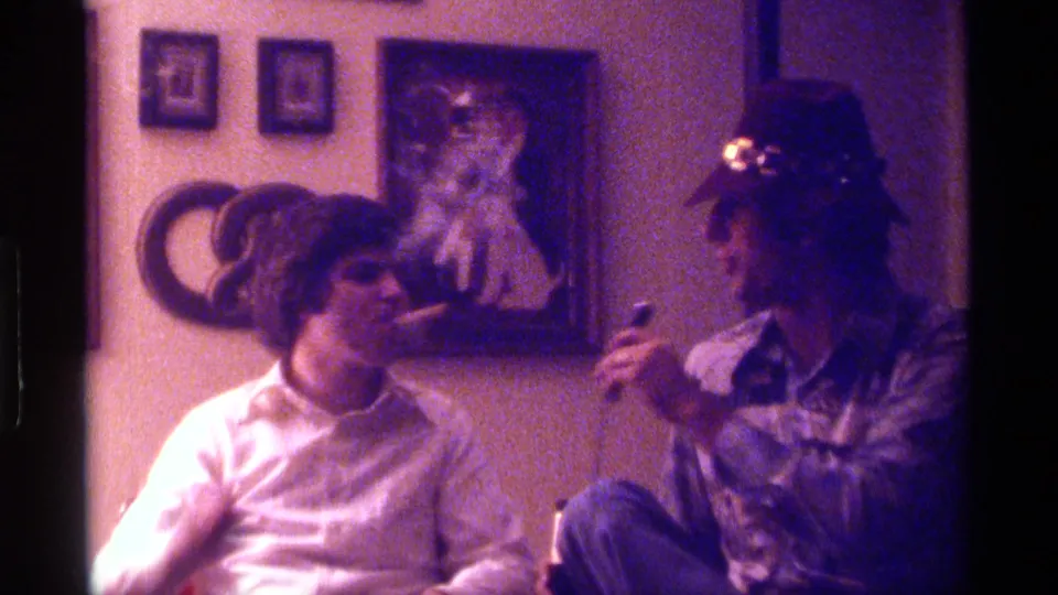 1976 Kentucky home-movie frame of a young man smoking beside a friend with a microphone during a casual indoor gathering.