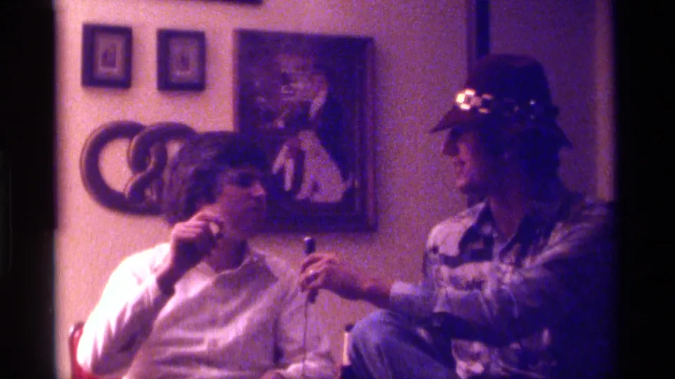 1976 Kentucky home-movie frame of two friends indoors, with one smoking and the other extending a microphone during an informal exchange.