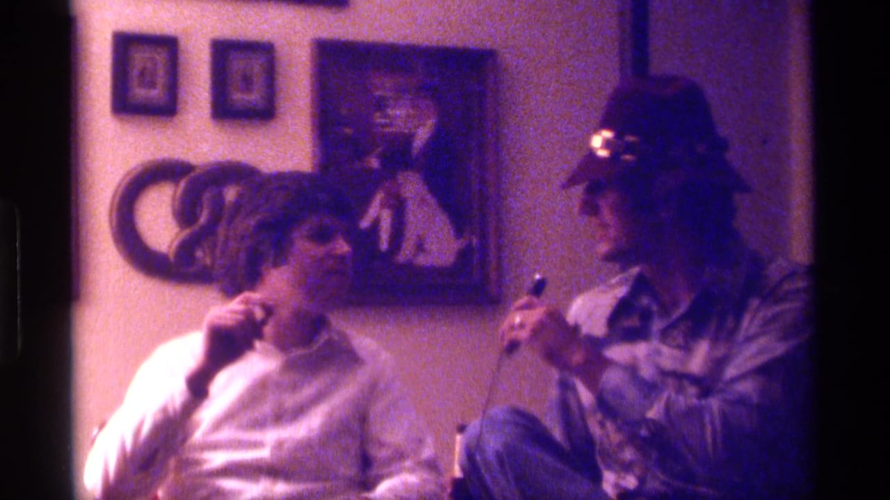 1976 Kentucky home-movie frame of two adult men indoors, one in a tan hat holding a microphone toward a seated man in a white shirt.