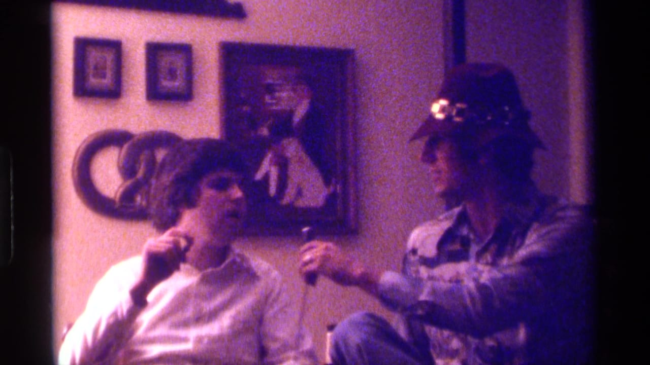 Man in tan hat extends a microphone toward a seated man in a white shirt indoors.