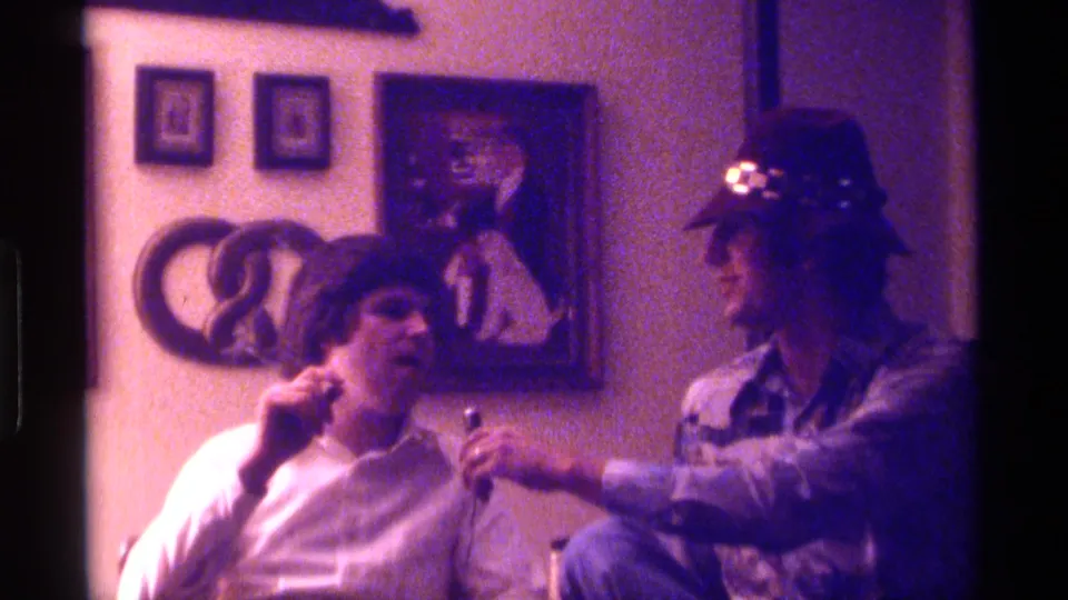 1976 Kentucky home-movie frame showing a seated man in a white shirt responding as a man in a tan hat holds a microphone nearby.