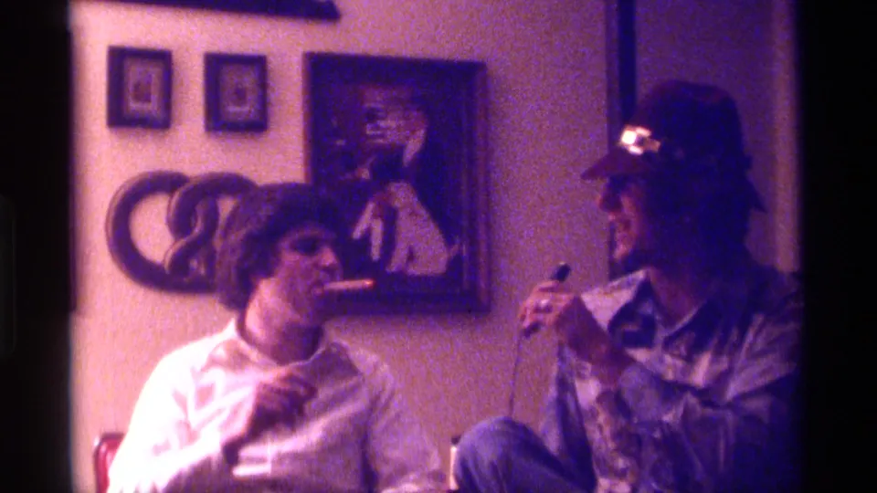 1976 Kentucky home-movie frame of two adult men seated indoors with a handheld microphone and wall art behind them.